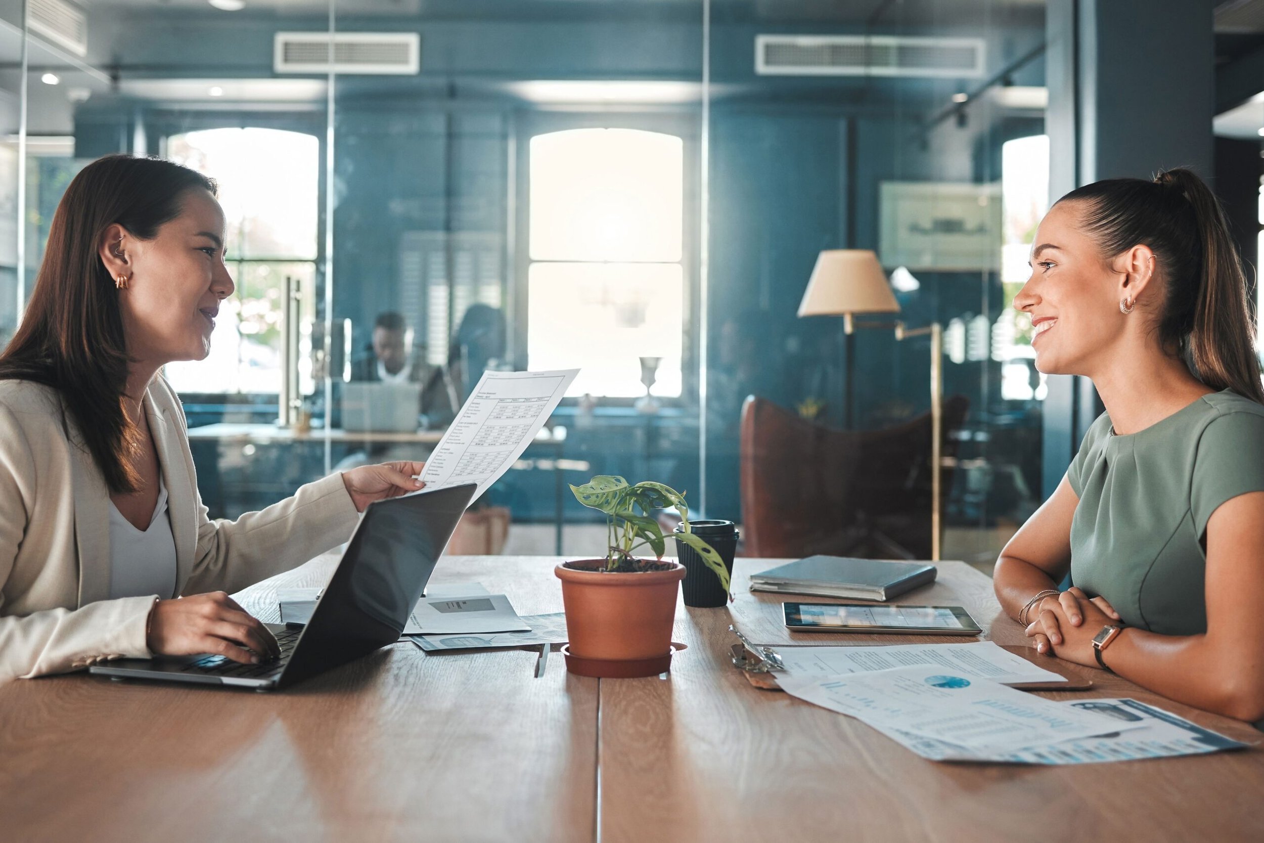 Two women sitting across from each other at a wooden table in a modern office, engaged in conversation. One woman, holding a document, appears to be explaining something to the other. The table has a potted plant, papers, and a digital tablet. The office features large windows and glass walls, with a person working in the background.
