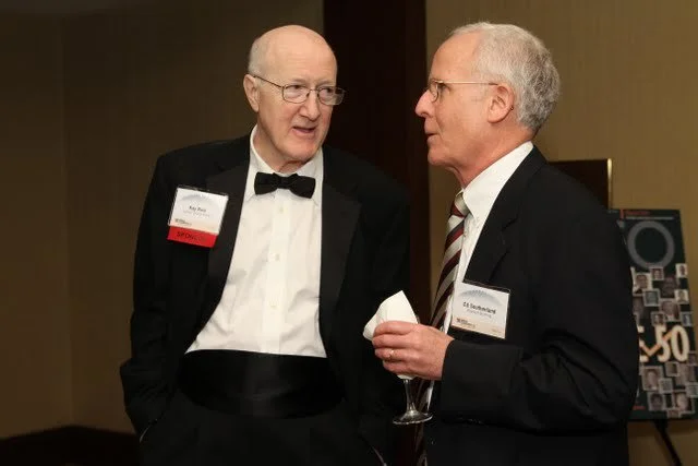 Two older men in tuxedos and glasses conversing at a formal event, one holding a glass of wine.