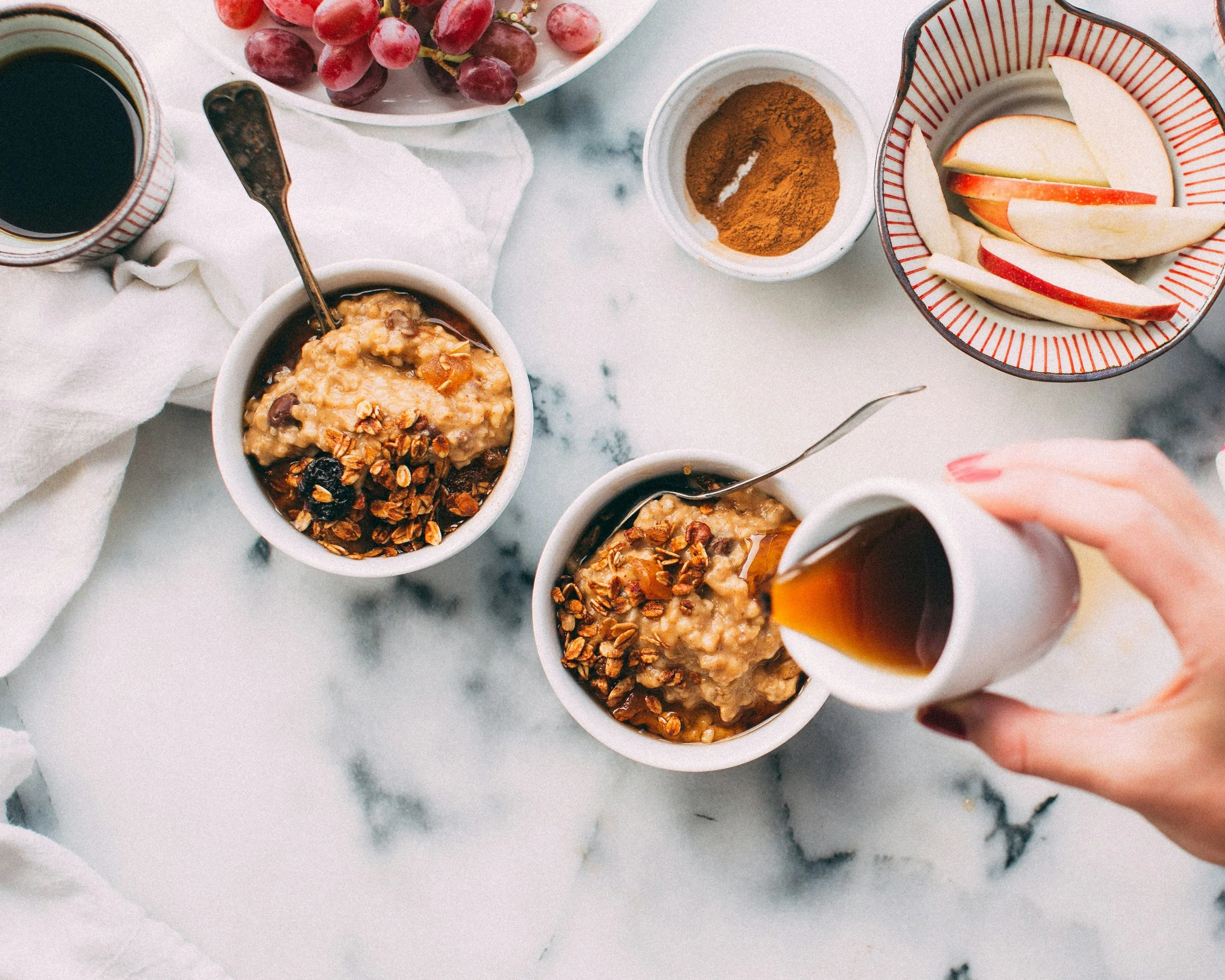 Raisin, pommes, épices et granola dans des petits bols, tasse de café, assiette de raisins rouges, petits bols de pommes tranchées et épices sur une table en marbre blanc.
