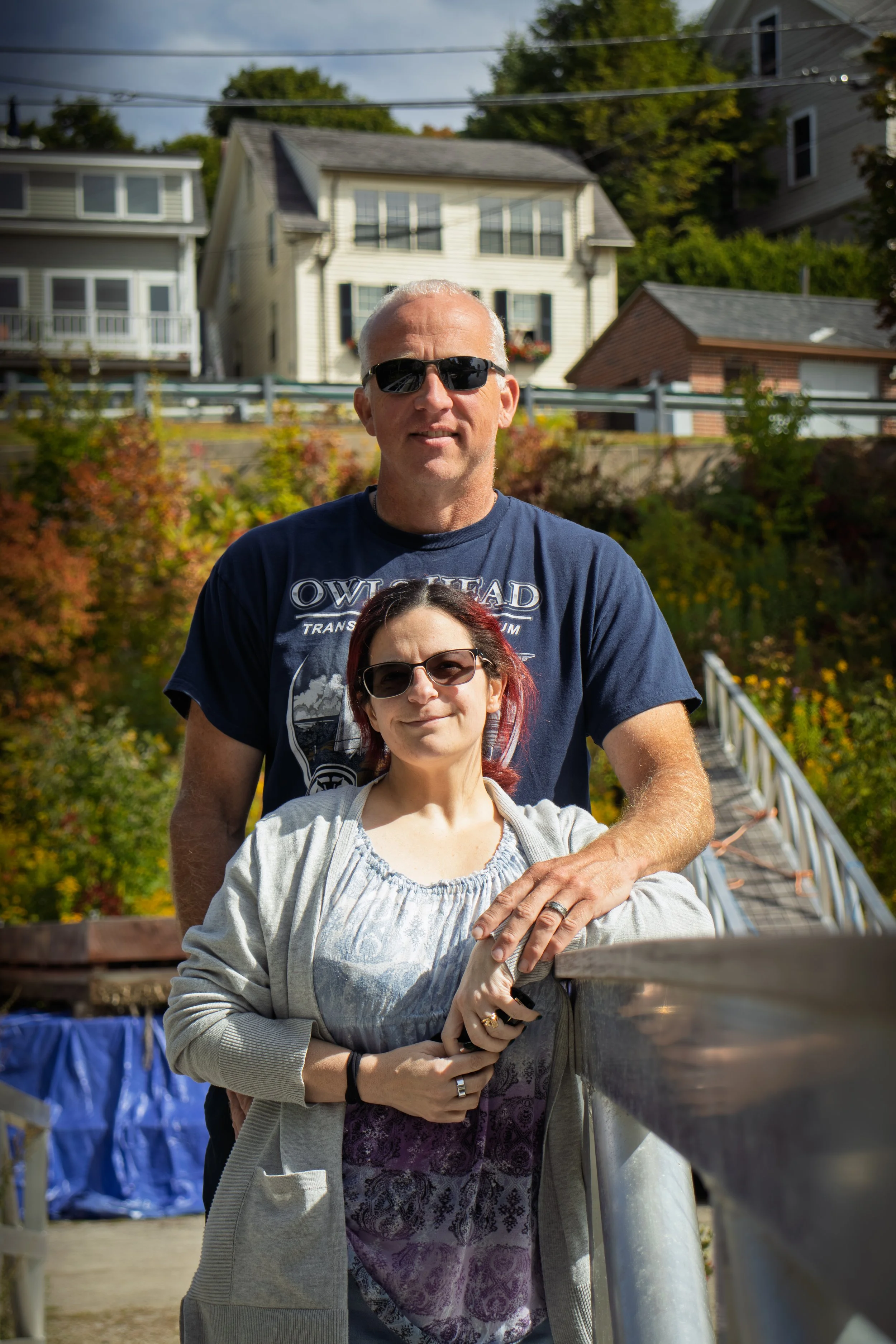 A man with white hair wearing sunglasses and a dark t-shirt standing behind a woman with glasses and red hair, on a bridge outdoors during daytime.