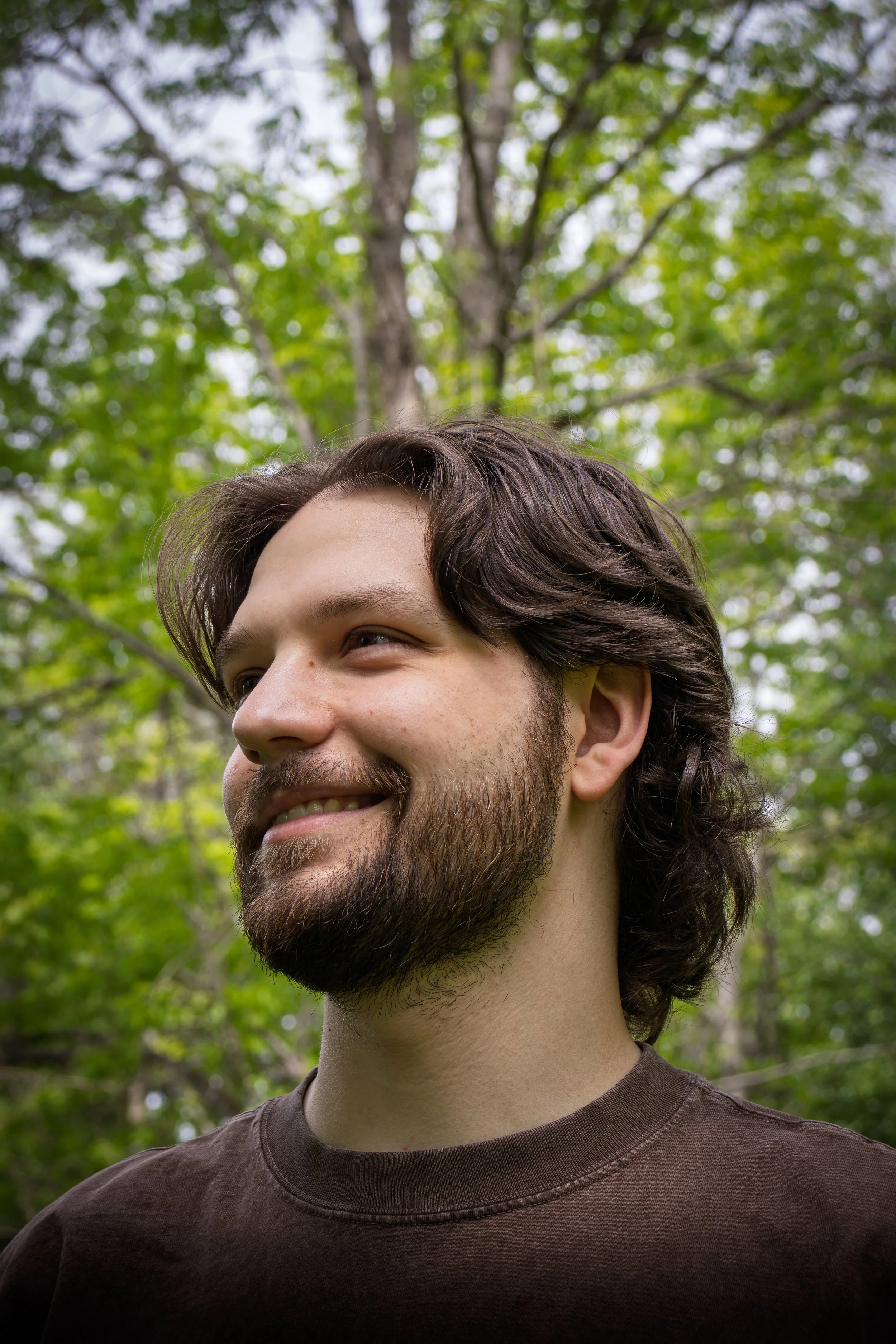 Close-up of a smiling young man with brown hair and a beard outdoors, with green trees and sky in the background.