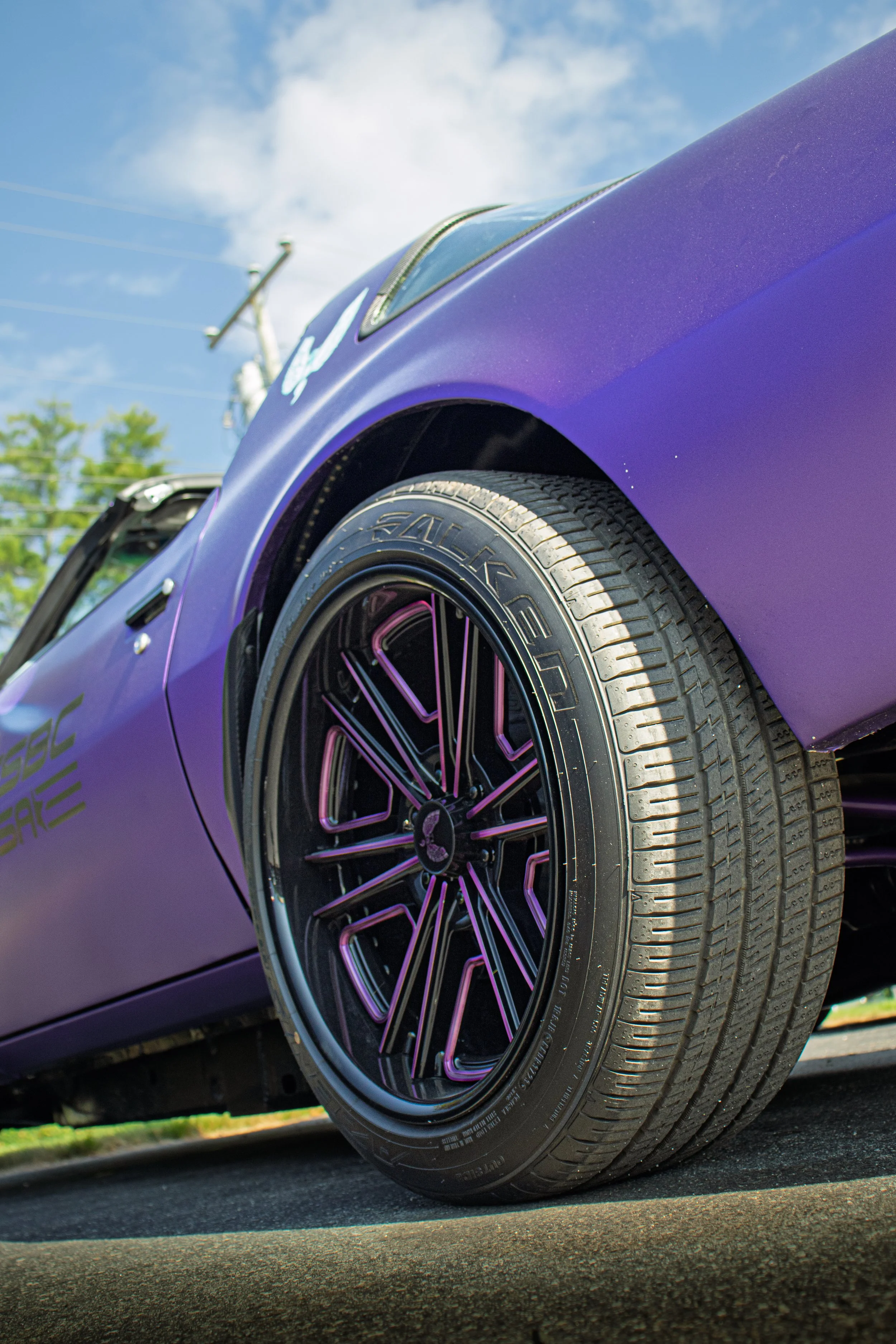 Close-up of a purple sports car's front tire and wheel, with a smooth tire and hot pink accents on the rim, set against a background of a blue sky with a few clouds and power lines.