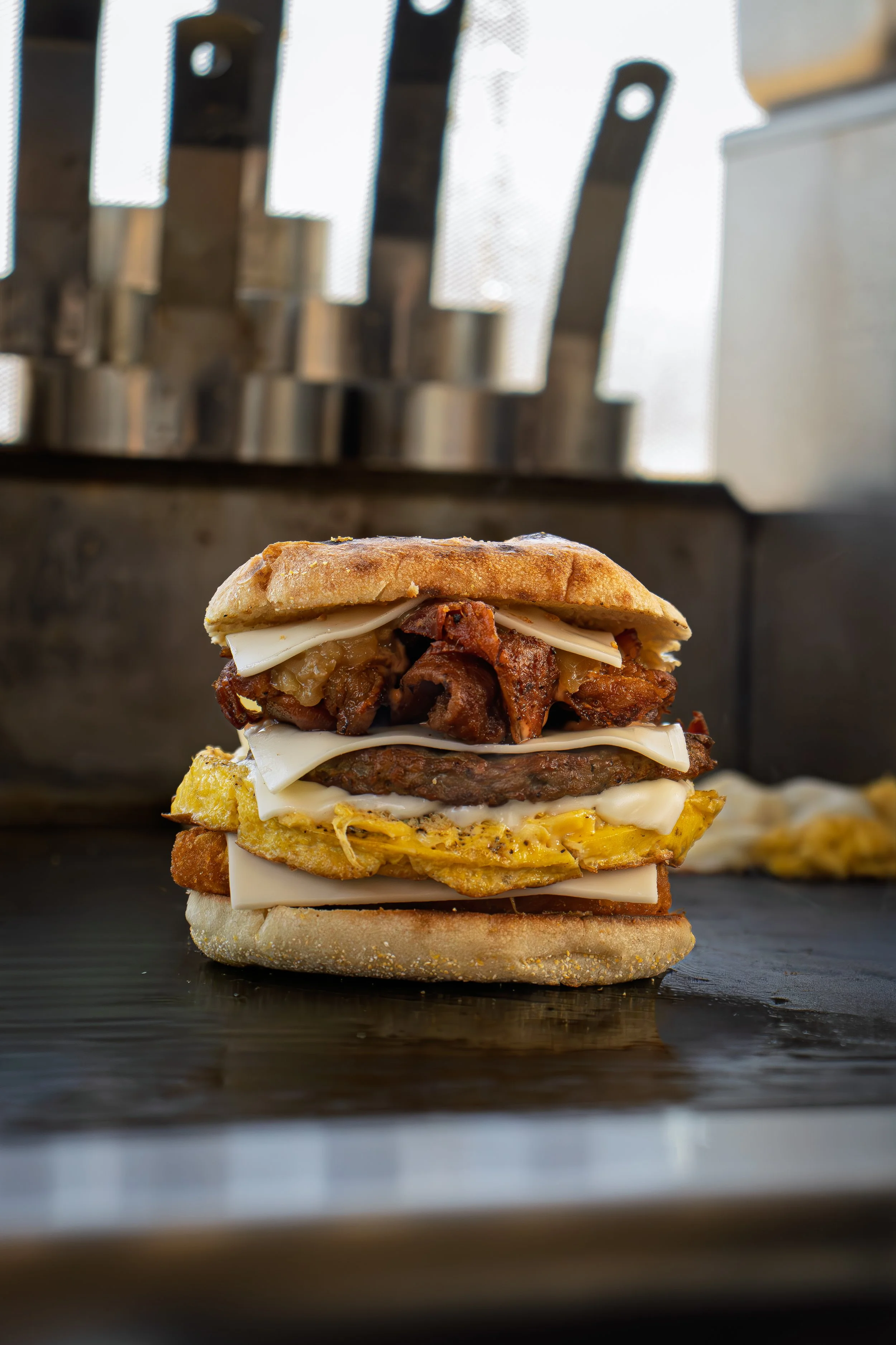 Close-up of a stacked burger with various meats and cheese, set against an industrial kitchen background.