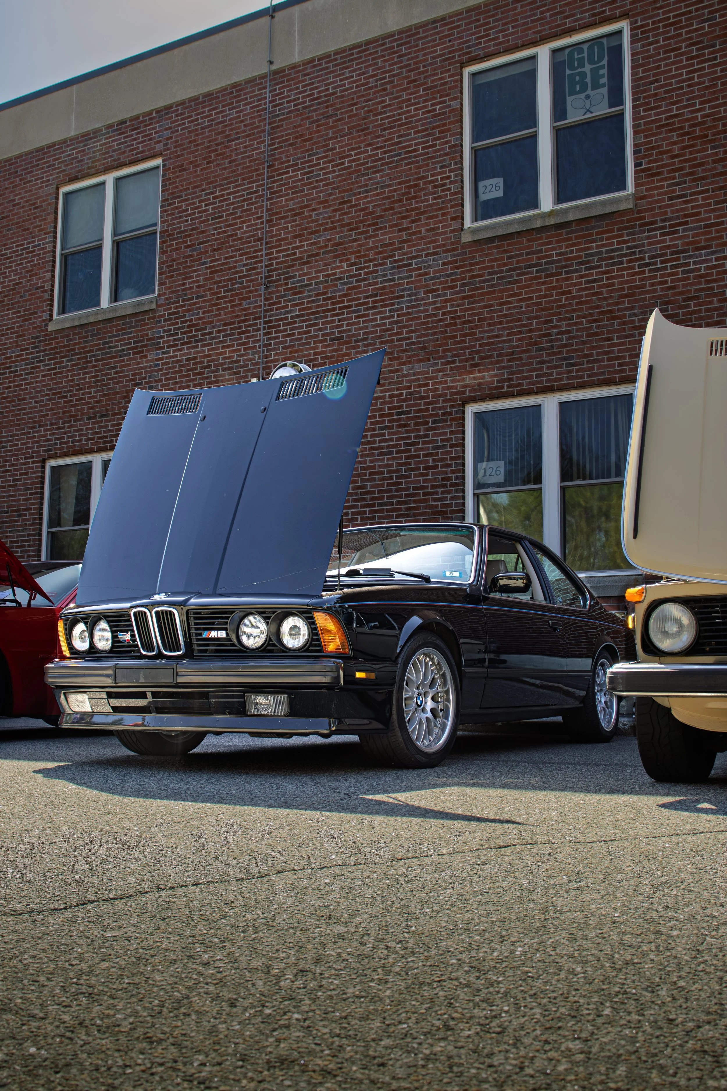 Several vintage cars, including a black BMW with its hood open, parked in a lot outside a brick building.