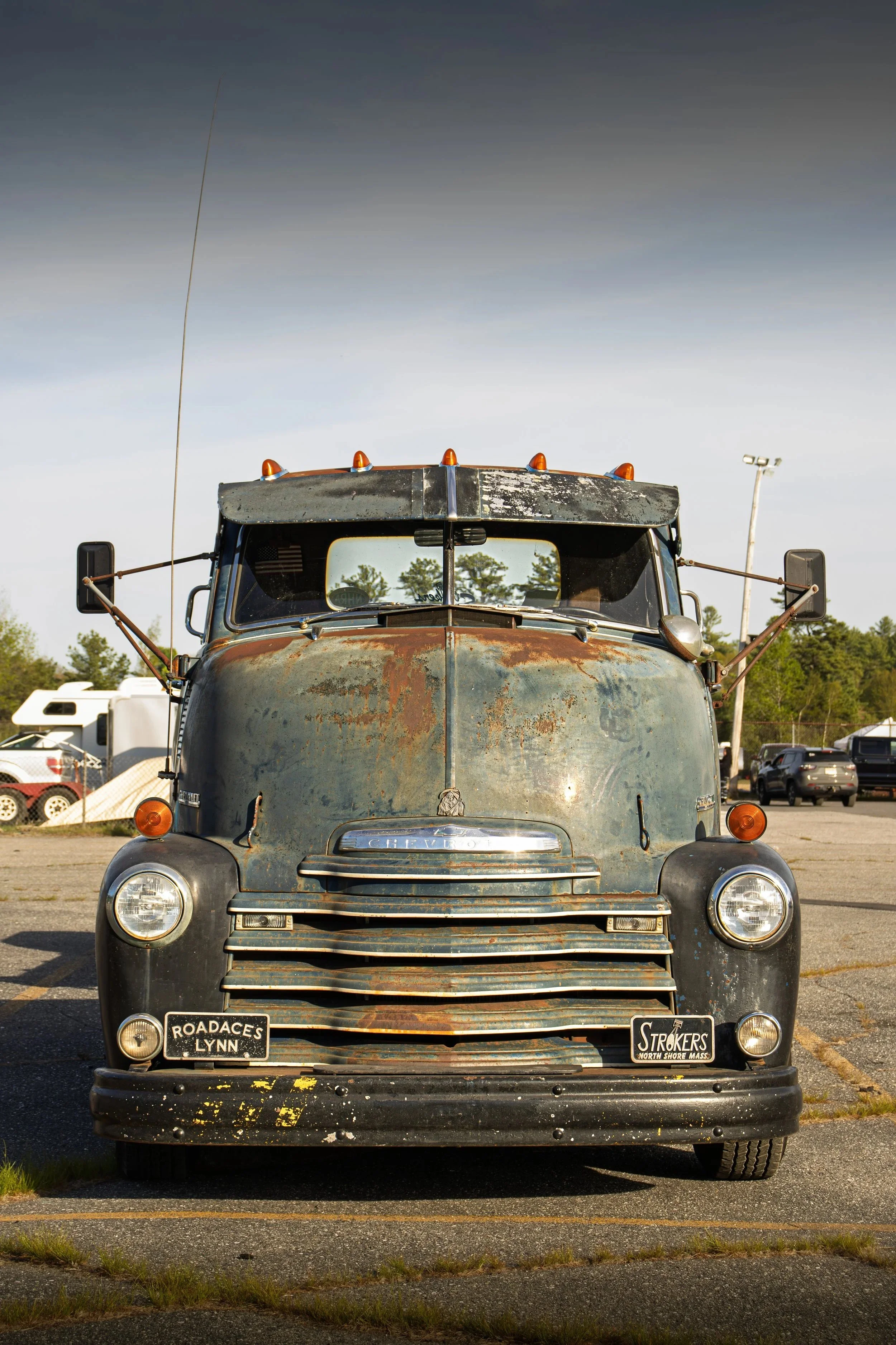 Front view of an old, rusty black truck parked in a lot, with a cloudy sky and trees in the background.
