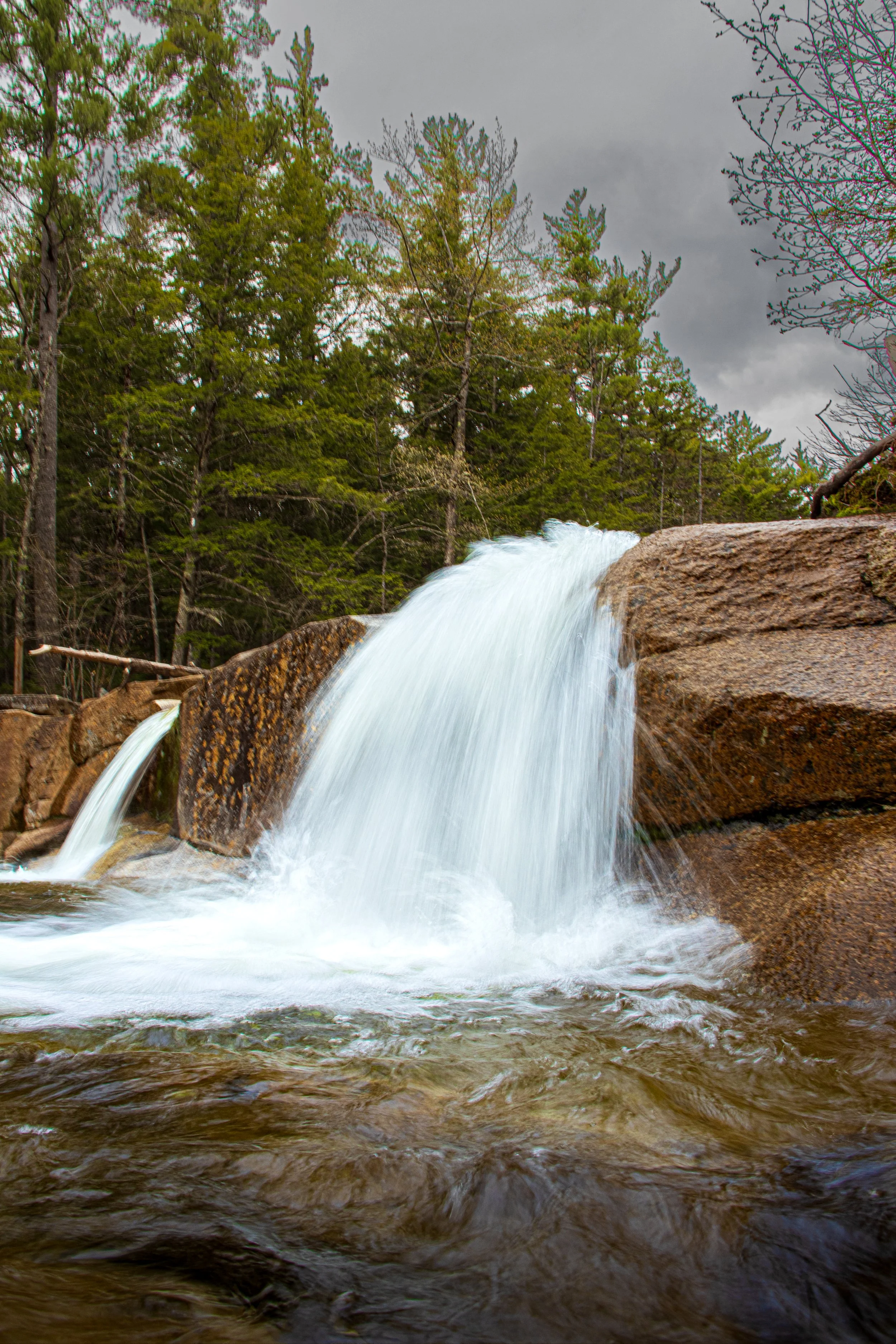A small waterfall flowing over rocks in a forested area with green trees and a cloudy sky in the background.