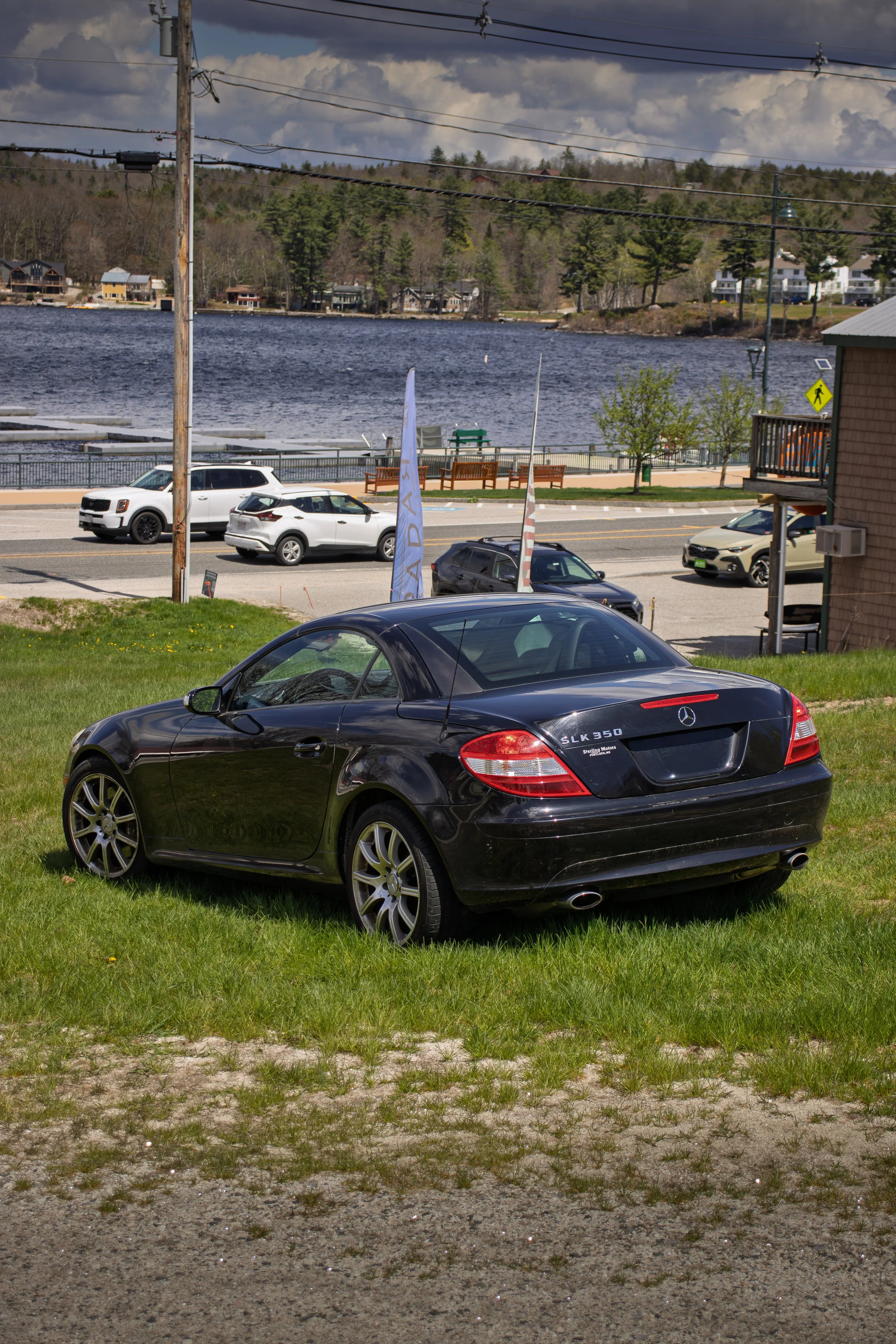 A black Mercedes-Benz SLK 350 convertible parked on a grassy area with a gravel patch in the foreground. In the background, a road with several cars drives past a waterfront with benches, trees, and a railing. Across the water, there are houses and a wooded hillside under a partly cloudy sky.