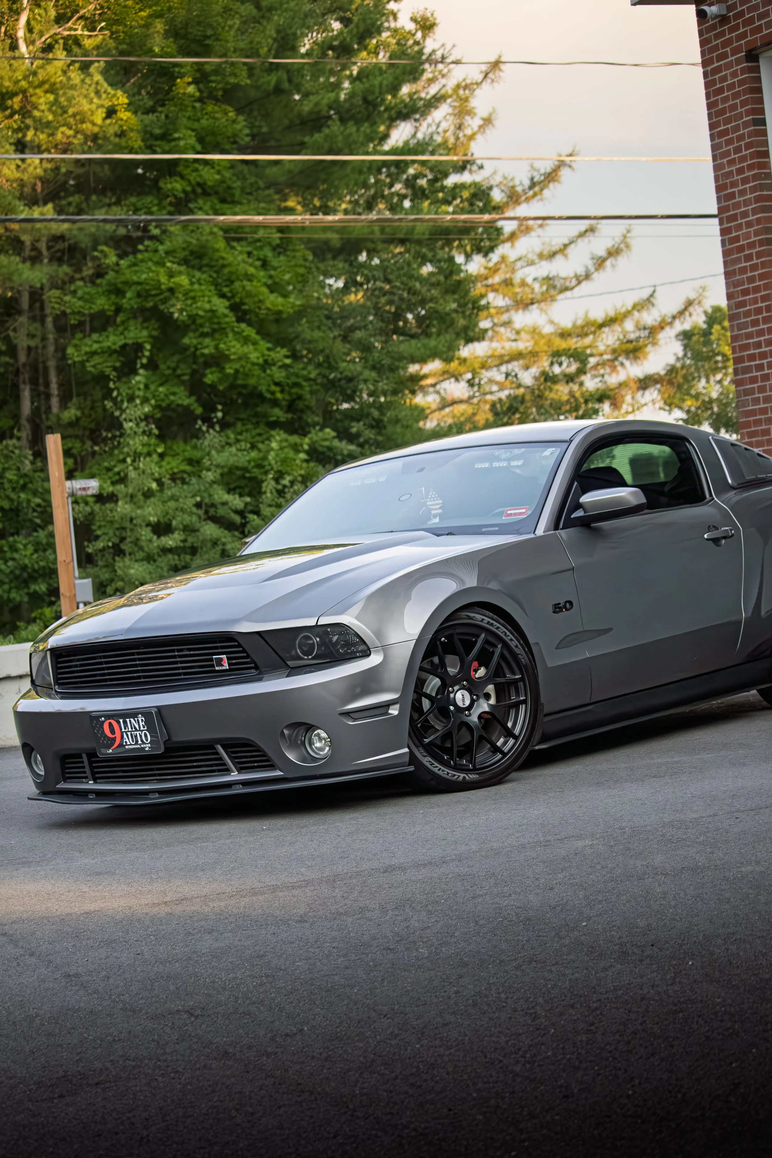 A silver Ford Mustang Roush parked on a paved driveway with green trees and a brick building in the background.