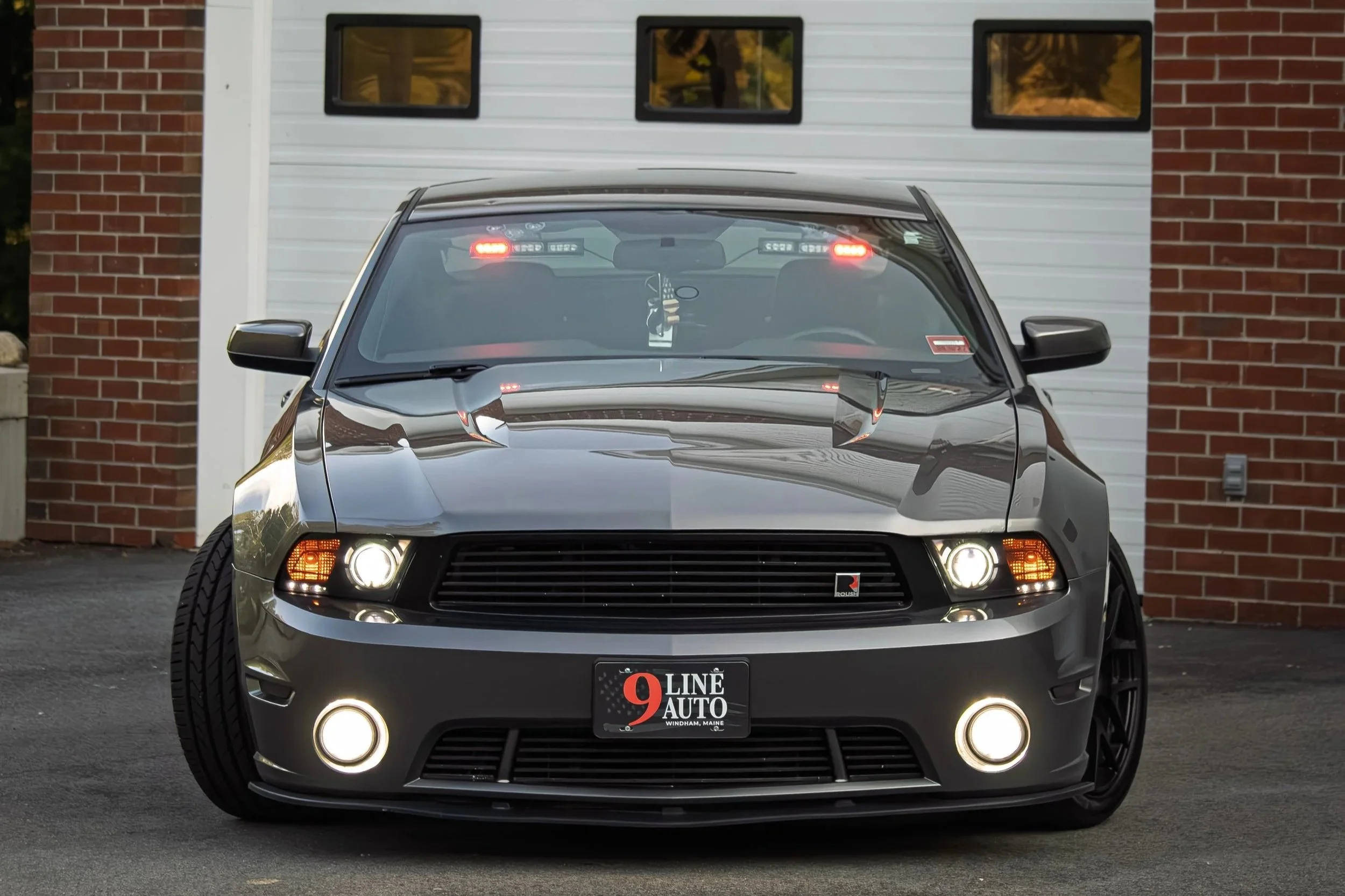 Front view of a mustang parked in front of a building with brick walls and white siding, showing the car's grille, headlights, and windshield with a rearview mirror and interpretation card hanging inside.