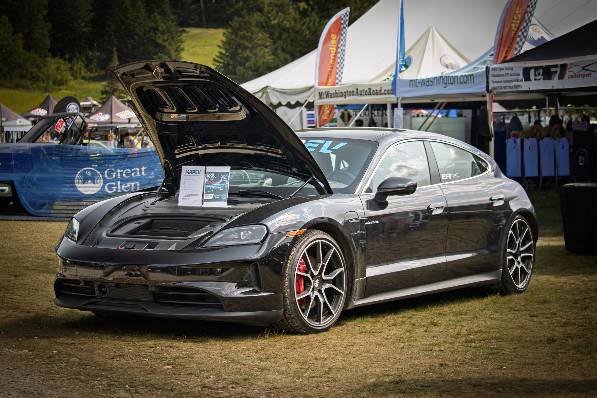 A black sports car with its hood open displayed at an outdoor auto event, tents and banners in the background.