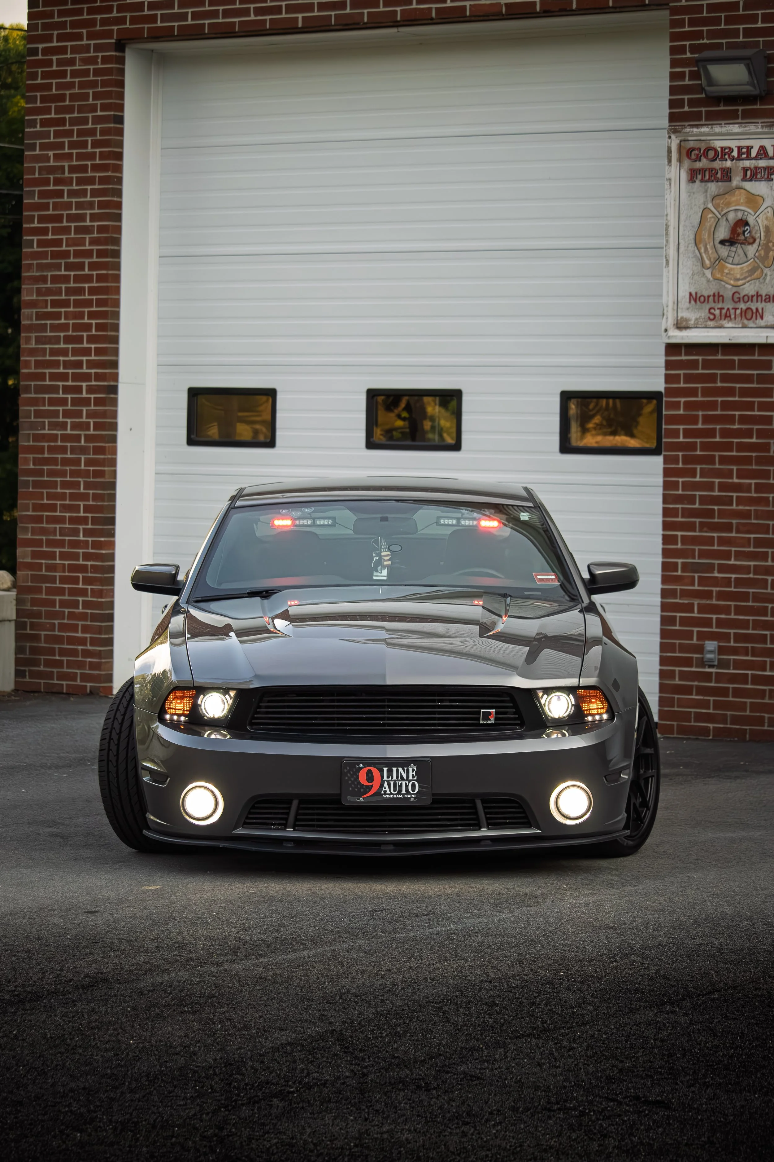 Front view of a gray Ford Mustang parked on black asphalt in front of a fire station with a brick wall and a white garage door with small windows.