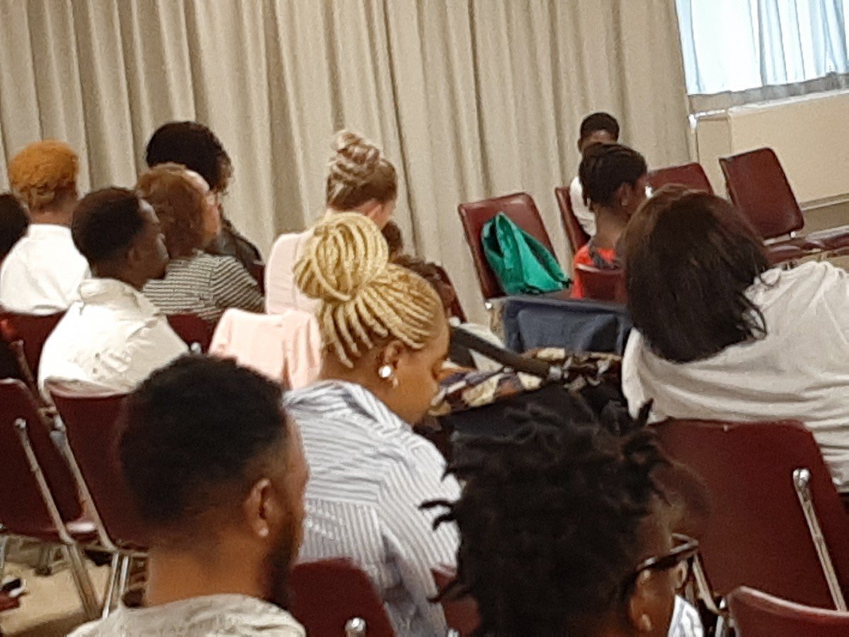 People seated in chairs attending a conference or seminar in a room with curtains and a window.