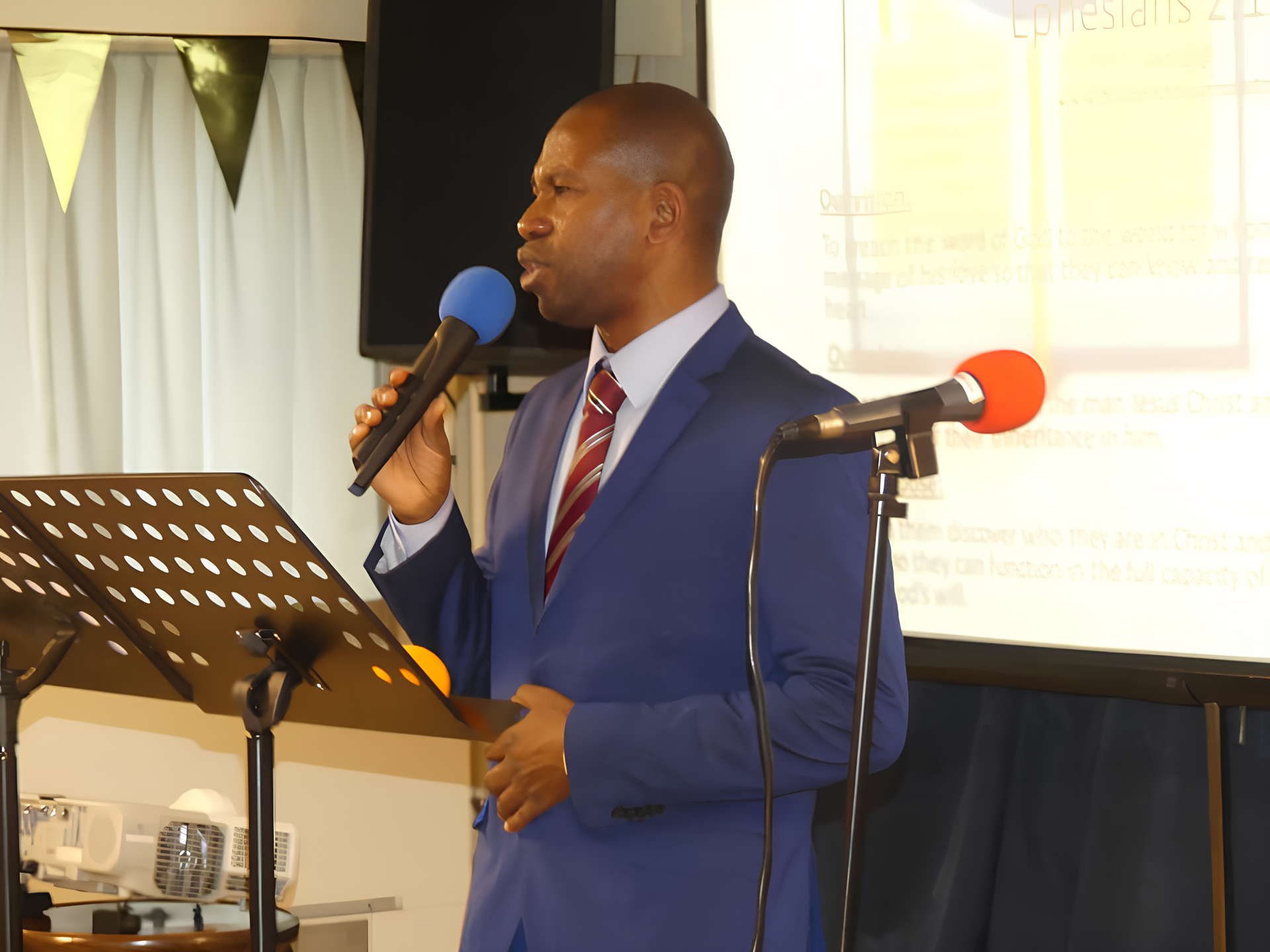 A man in a blue suit, white shirt, and red striped tie speaking into a blue microphone at a presentation or event. There is a music stand in front of him and a whiteboard in the background with some partially visible text and a large screen or monitor.