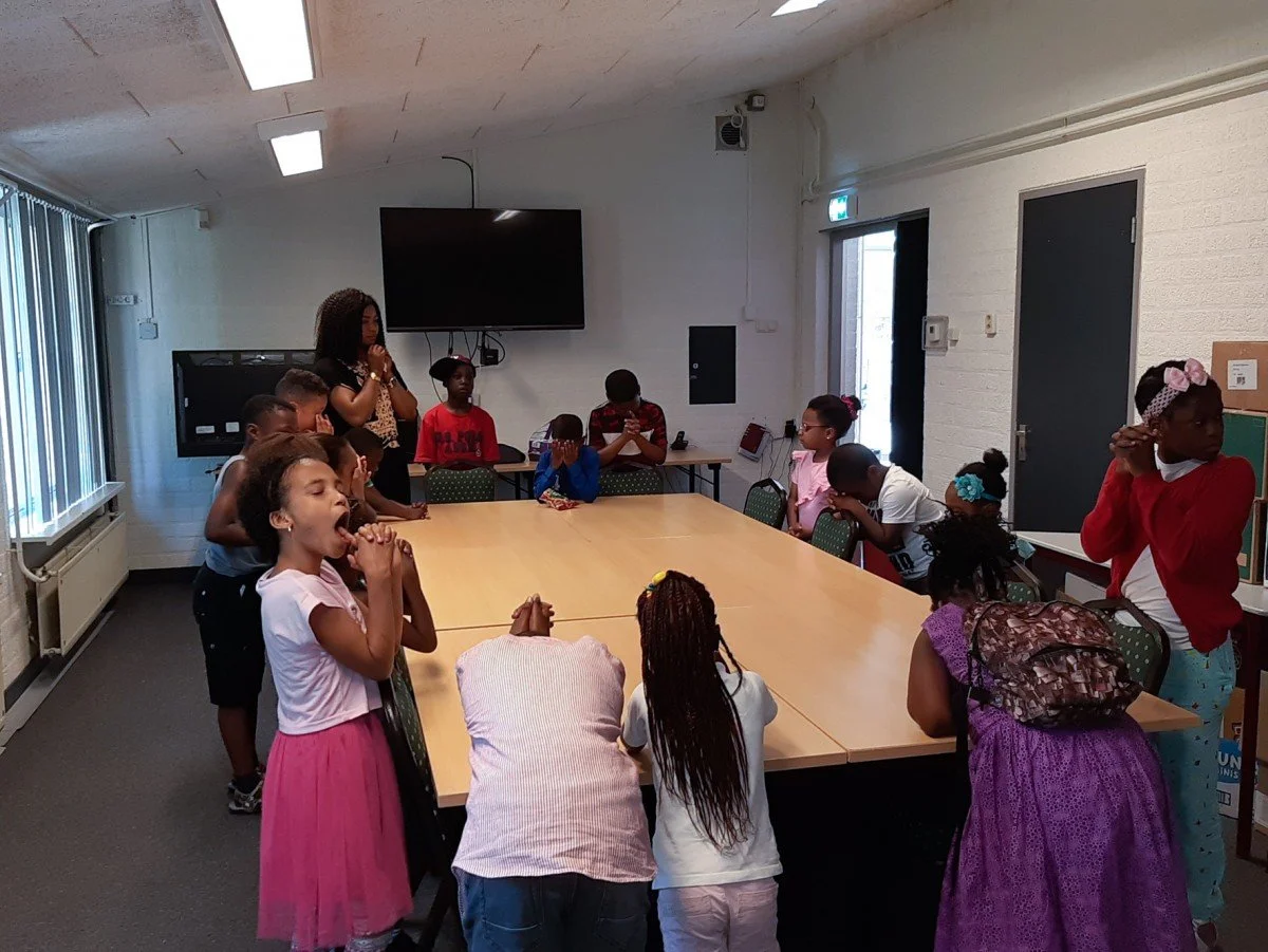 Children and a woman gathered around a large rectangular table in a classroom or meeting room, some with eyes closed and hands clasped or raised, possibly praying, with a large screen on the wall behind them.