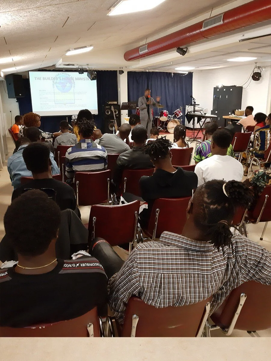 A group of people seated in a church or event hall, listening to a speaker at a podium on stage with musical instruments and a large screen displaying a presentation.