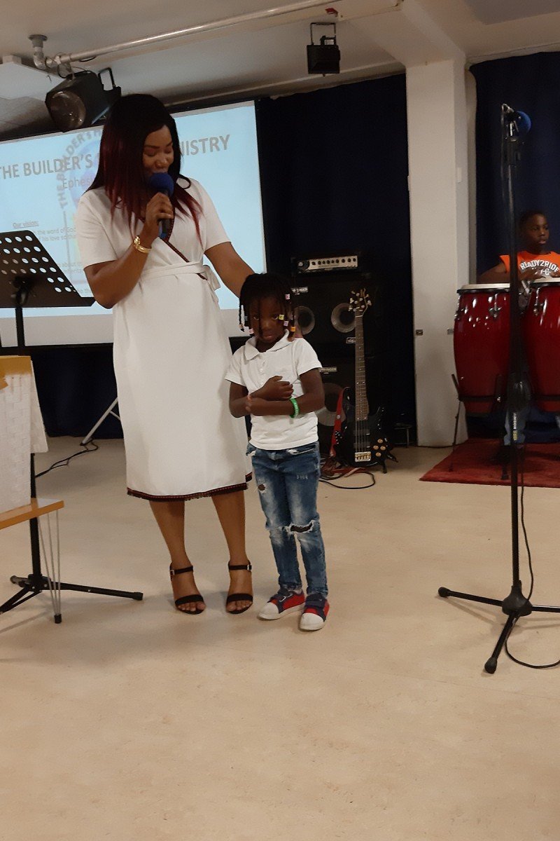 A woman in a white dress is speaking into a microphone on stage with a child standing beside her. The child has glasses and is dressed casually. There are musical instruments and a projector screen in the background.