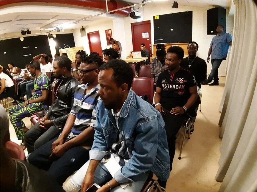 Group of people sitting on chairs in a conference room or event space, attentively listening, with some standing in the back, and a woman standing near the wall.