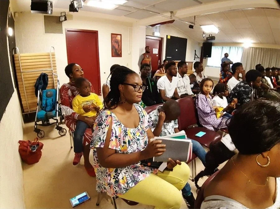 Group of people seated in a room, attending a presentation or class, with some taking notes and children present.
