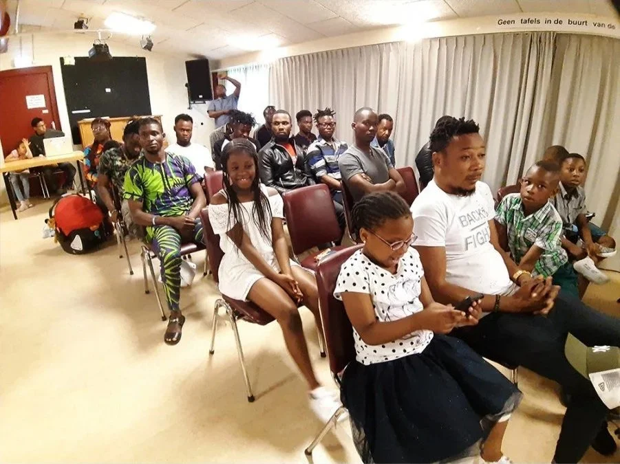 Group of children and adults sitting on chairs in a room with beige walls and curtains, some looking at their phones.
