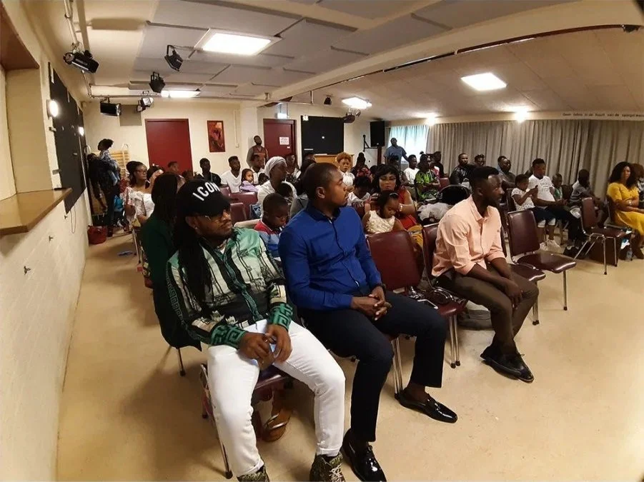 A group of people seated in a room, attending what appears to be a presentation or meeting. The room has a low ceiling, curtains on the windows, and some wall decorations.