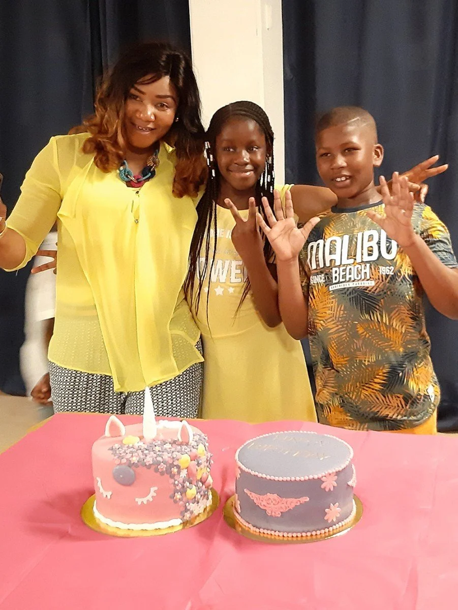 A woman and two children standing behind a table with two decorated birthday cakes, celebrating a birthday. The woman is wearing a yellow blouse, the girl in middle is wearing a yellow dress, and the boy on the right is wearing a tropical print shirt