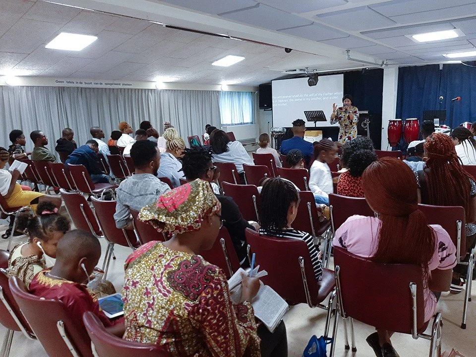 A diverse group of people attending a presentation or lecture in a conference room, with a speaker at the front. Some people are taking notes or reading a book, and there are musical instruments like drums behind the speaker.