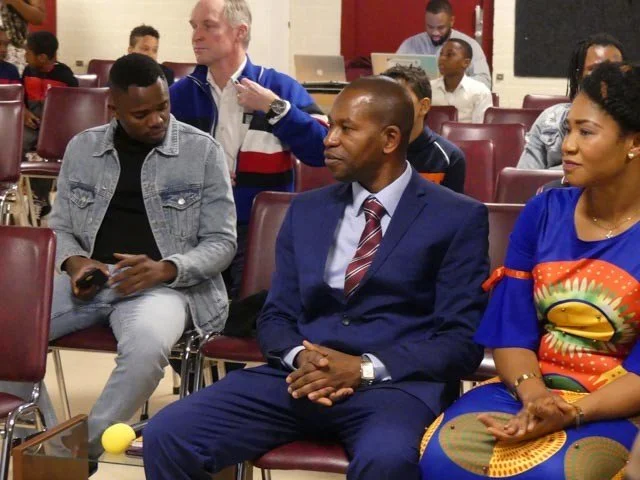 People seated in a room, one man in a suit and tie, a woman dressed in colorful traditional attire, and others in casual clothing, in what appears to be a meeting or event.