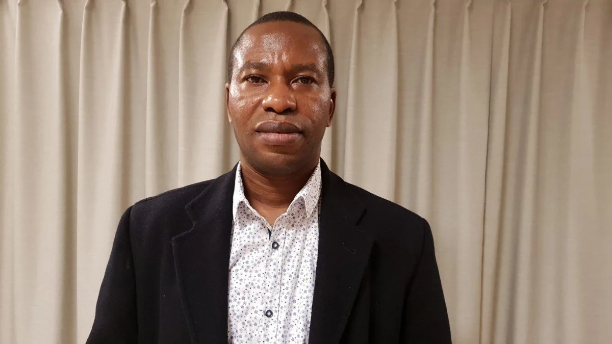 A man in a black blazer and white collared shirt standing in front of beige curtains.