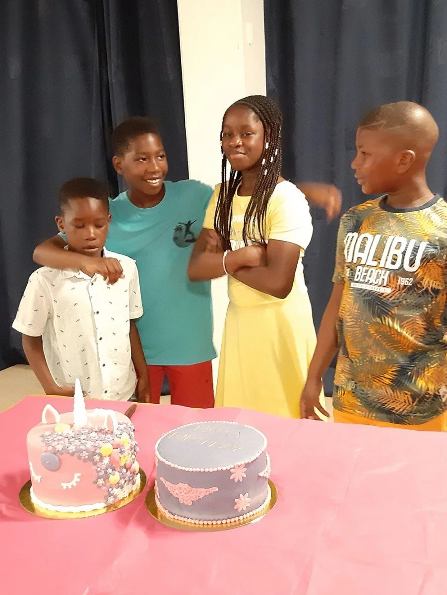 Children celebrating a birthday with two decorated cakes on a pink tablecloth.