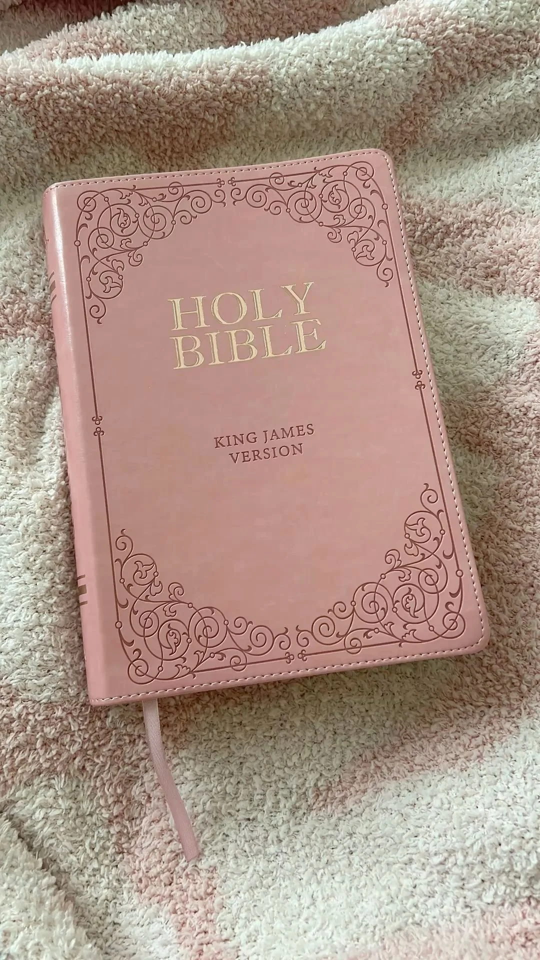 A pink book cover of the Holy Bible with ornate border design, labeled 'King James Version', resting on a soft, fluffy, beige and pink blanket.