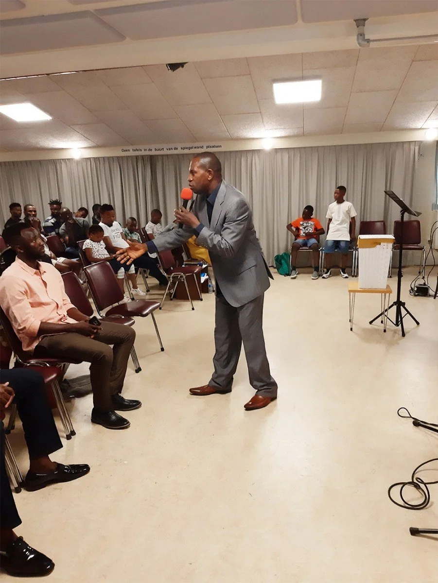 A man in a gray suit speaks into a microphone during an indoor event, gesturing with his hand, with a diverse audience seated and standing around him.