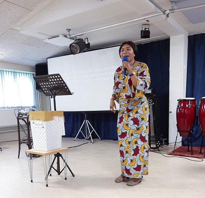 A woman in colorful floral clothing is speaking into a microphone on a stage with musical instruments, including conga drums, in the background.