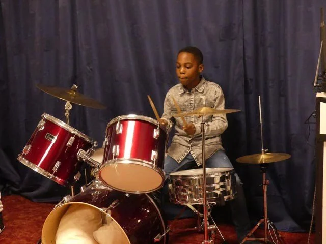 A young boy sitting behind a red drum set, holding drumsticks, with a dark backdrop and a music stand nearby.