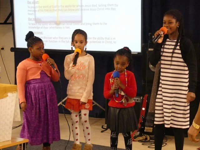 Four girls are holding microphones during a presentation or performance, with a woman playing a guitar and a projection screen in the background.