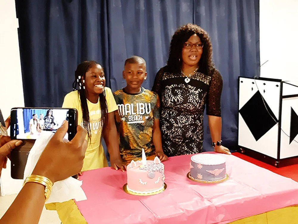 A woman and two children are celebrating a birthday, standing behind two decorated cakes on a pink tablecloth, with a person taking a photo.