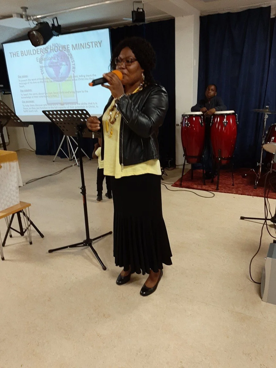 A woman singing into a microphone on a stage with a music band in the background during a church service or event, with a projection screen displaying a church-themed presentation.
