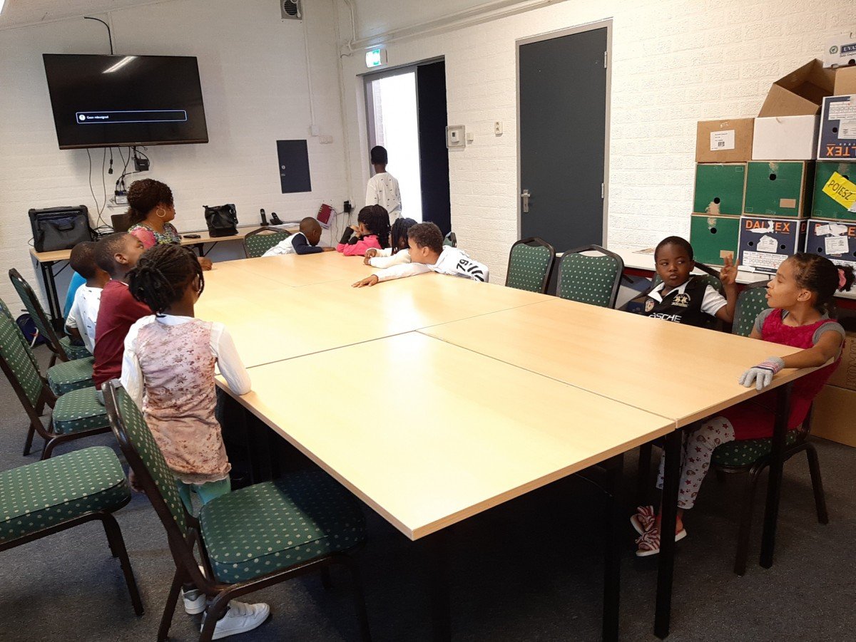 Children sitting around a large rectangular table in a classroom or meeting room, with one child standing near the door, a large wall-mounted TV, and boxes on a shelf in the background.