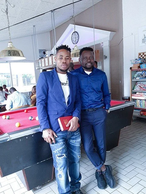 Two men dressed in blue suits standing next to each other in a pool hall, with other people sitting at tables in the background.