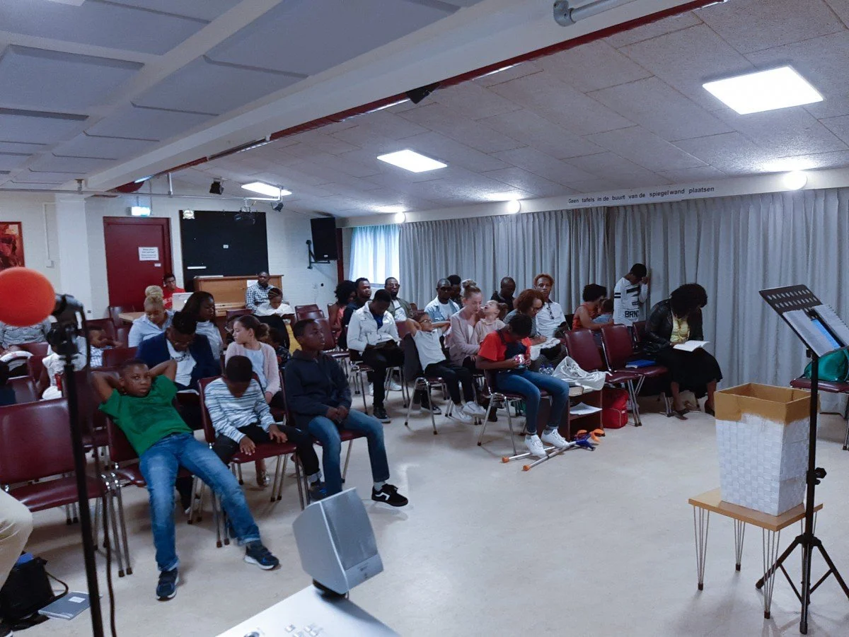 A group of children and adults seated in a room, likely at an event or gathering, with some looking at scripts or programs, and a music stand and trash bin visible in the foreground.