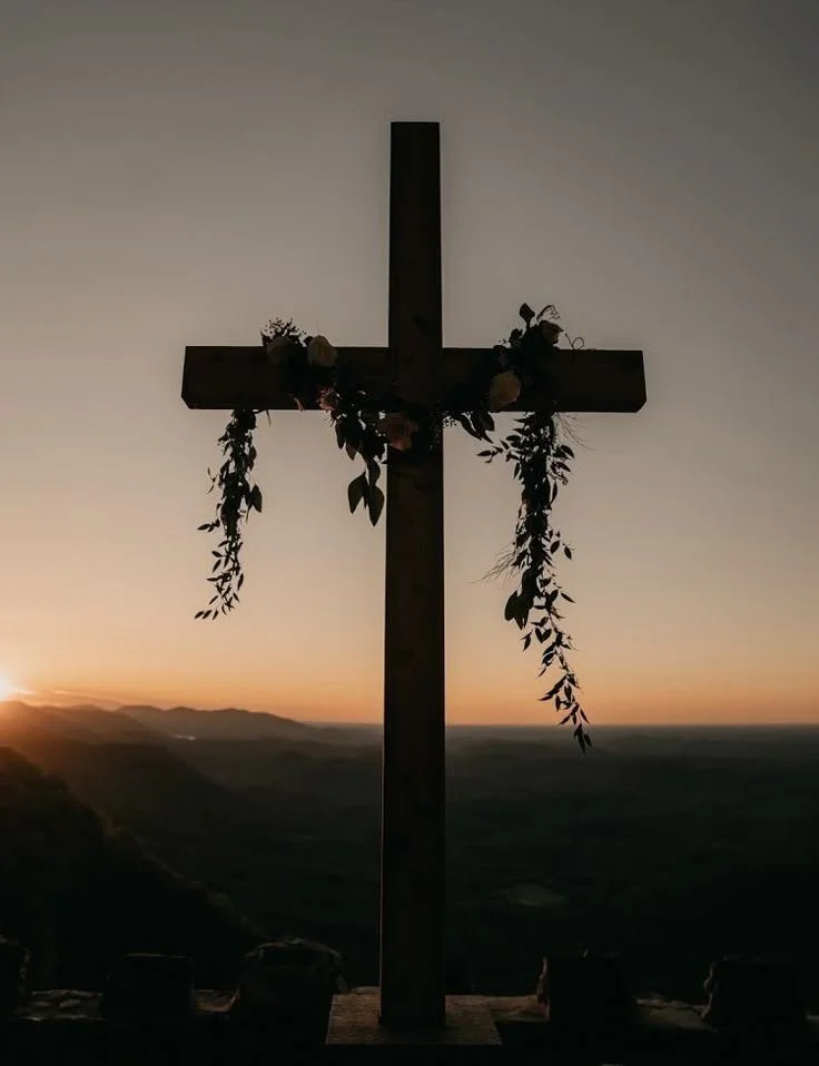 Wooden cross decorated with flowers and greenery, silhouetted against a sunset over a mountainous landscape.