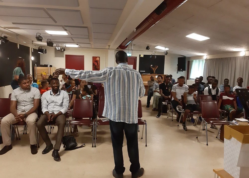 A man standing in front of a seated audience in a room, possibly giving a presentation or speech.