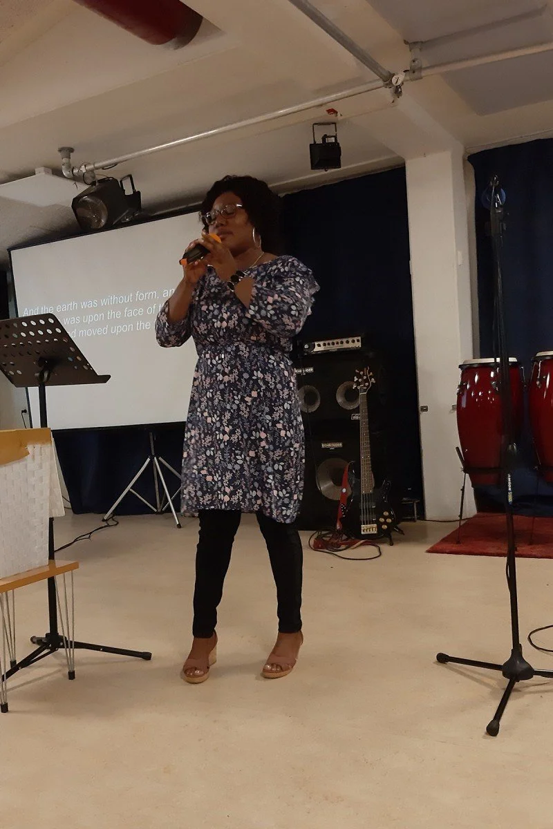 A woman singing into a microphone in a performance space with musical instruments and a presentation screen in the background.