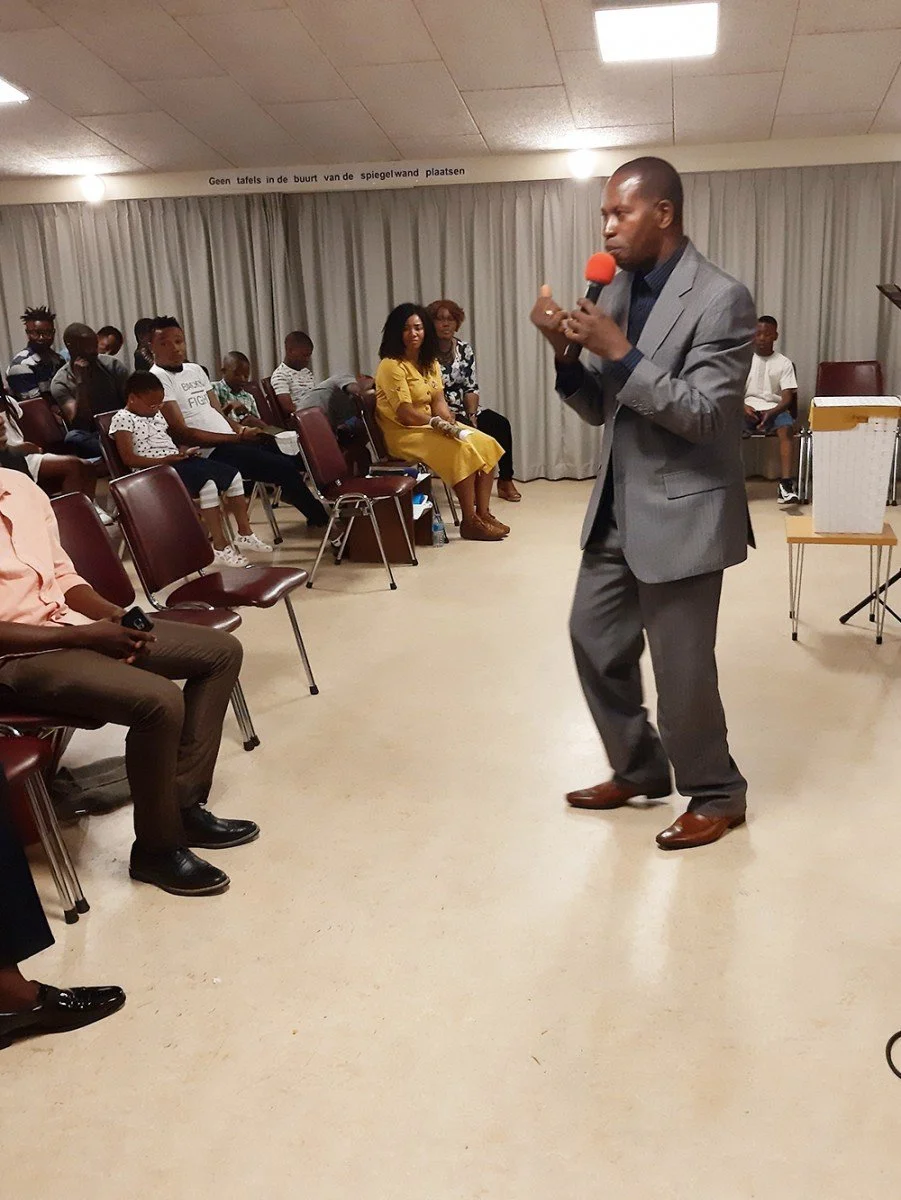 A man in a gray suit speaks into a red microphone during a presentation or speech in a room with seated audience members, including children and adults.
