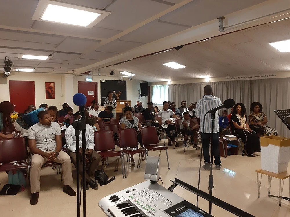 A group of people attending a presentation or seminar in a conference room, with a person speaking at the front, a microphone, and musical equipment in the foreground.
