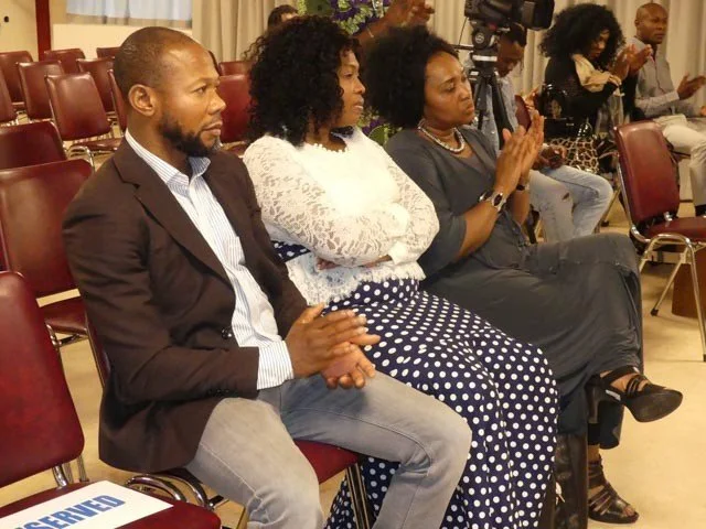 Three women and one man sitting in chairs at a formal event, with cameras and other people in the background.