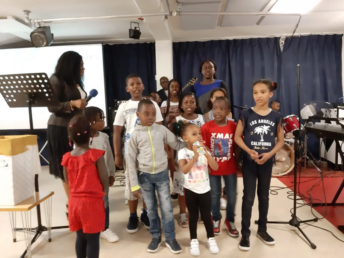 Group of children singing or performing on stage during a school or community event, with music equipment and a woman with a microphone.