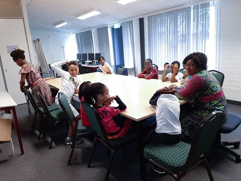 A group of children and an adult woman sitting around a large rectangular table in a classroom or meeting room, with some computers and curtains in the background.