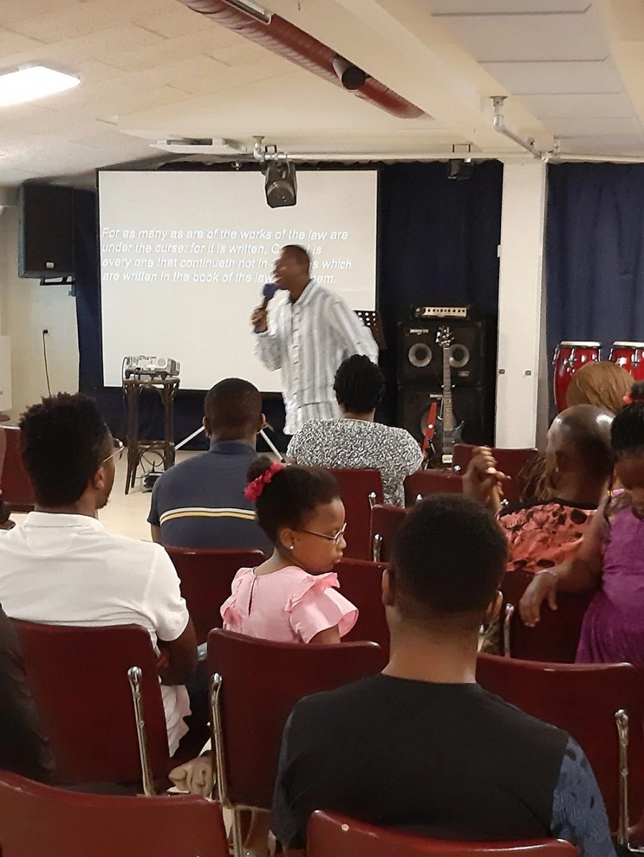 A man in striped shirt holding a microphone speaking to an audience of men, women, and children in a room with chairs, a projector screen, and musical instruments.