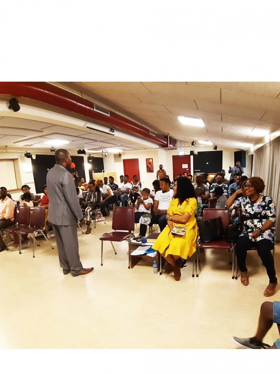 A man in a gray suit speaking to an audience in a conference room with many people sitting and listening. Some are taking notes or looking at their phones. Two women are seated in front, one in a yellow dress, the other in a floral top.