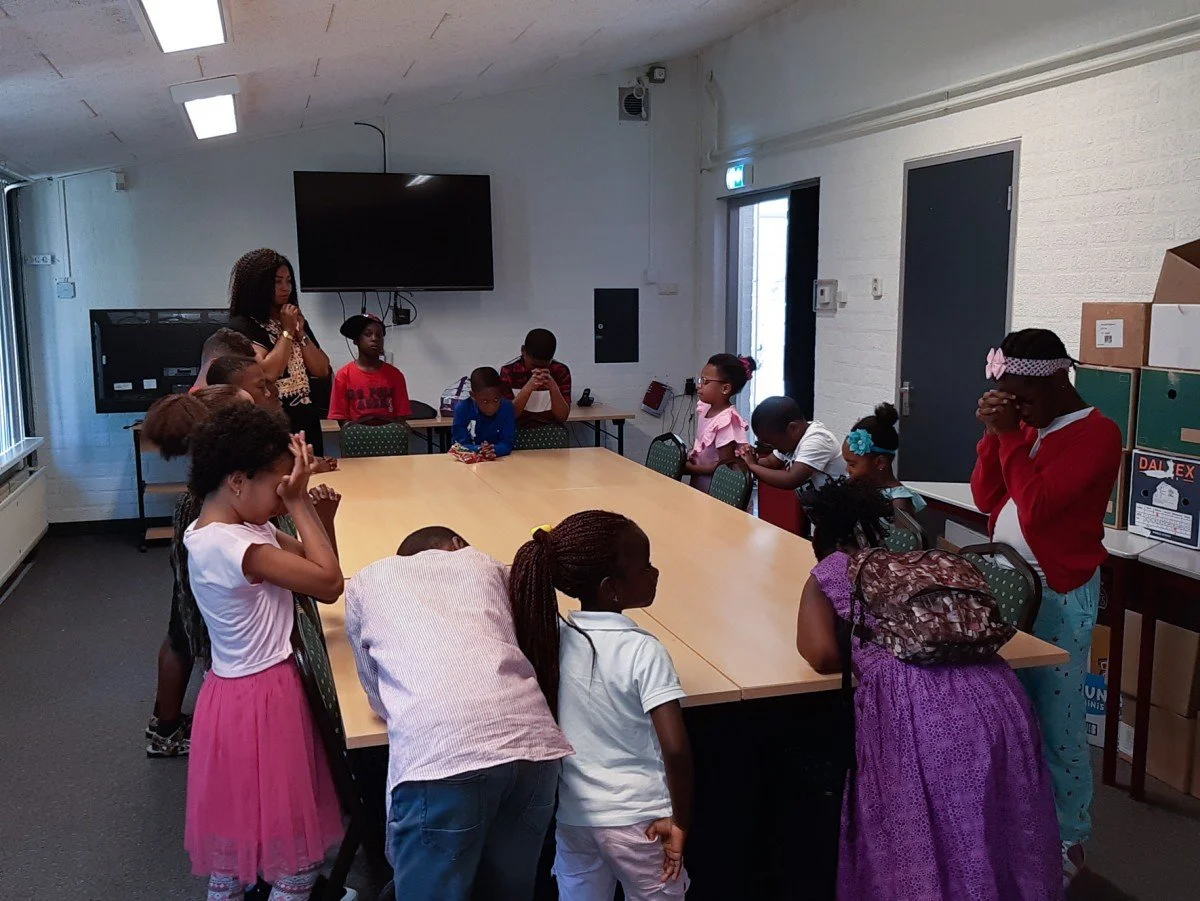 Children and a woman praying around a rectangular table in a room with white walls and a large TV on the wall.